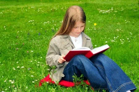 Adorable young kid reading a book while sitting on grass at the park